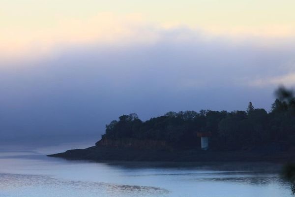 A serene landscape featuring a misty shoreline, trees, and a small structure on a calm body of water. The sky has soft colors.