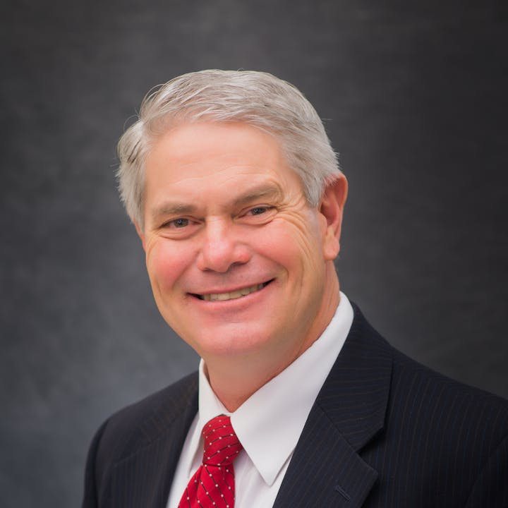 Board Member, Leonard Arave, in a suit and red tie with a gray background.