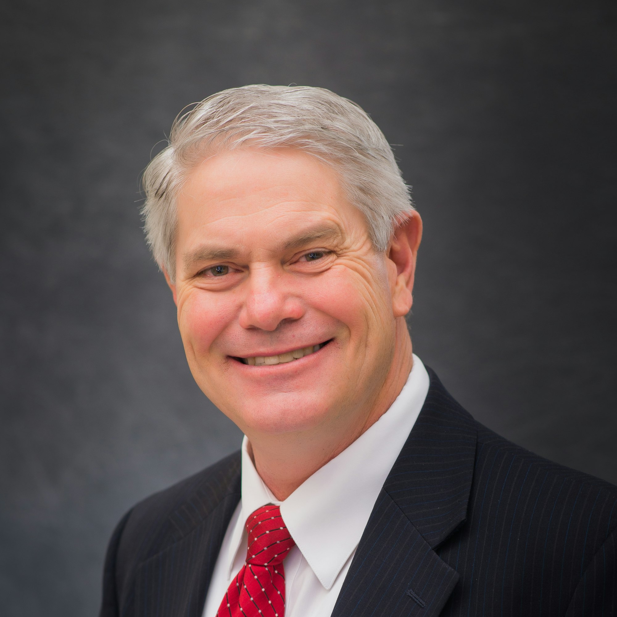 Board Member, Leonard Arave, in a suit and red tie with a gray background.