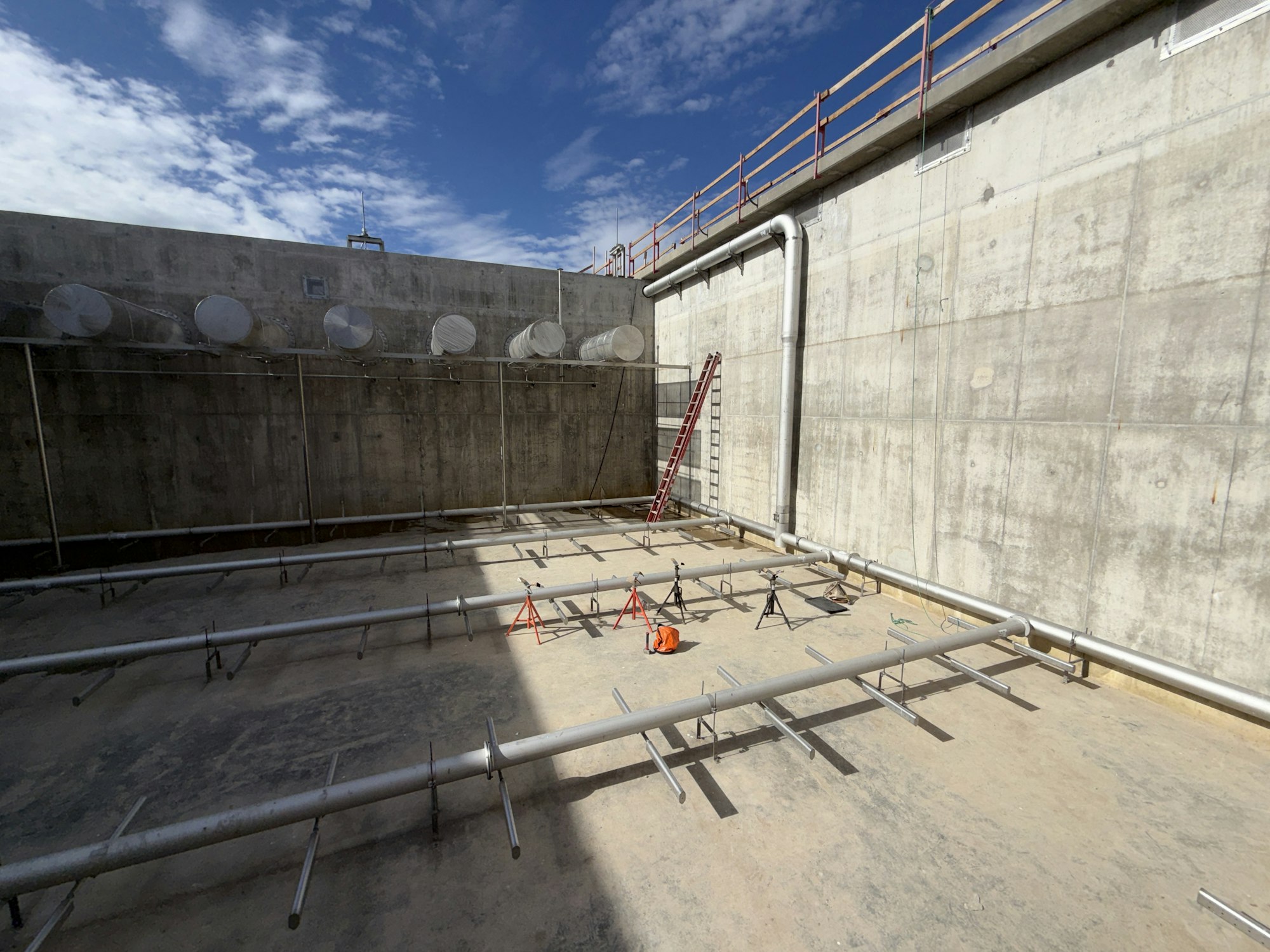 The image shows a large, concrete space with metal piping and a ladder, likely part of a construction or industrial site.