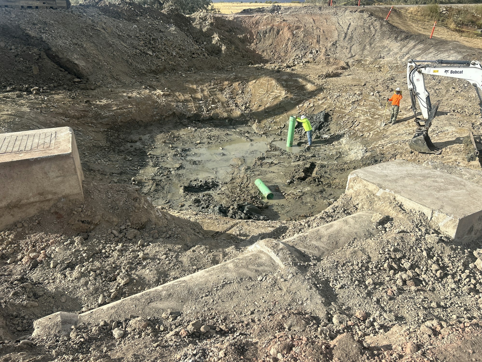 Construction site with workers installing green pipes in a large excavation filled with mud and debris.