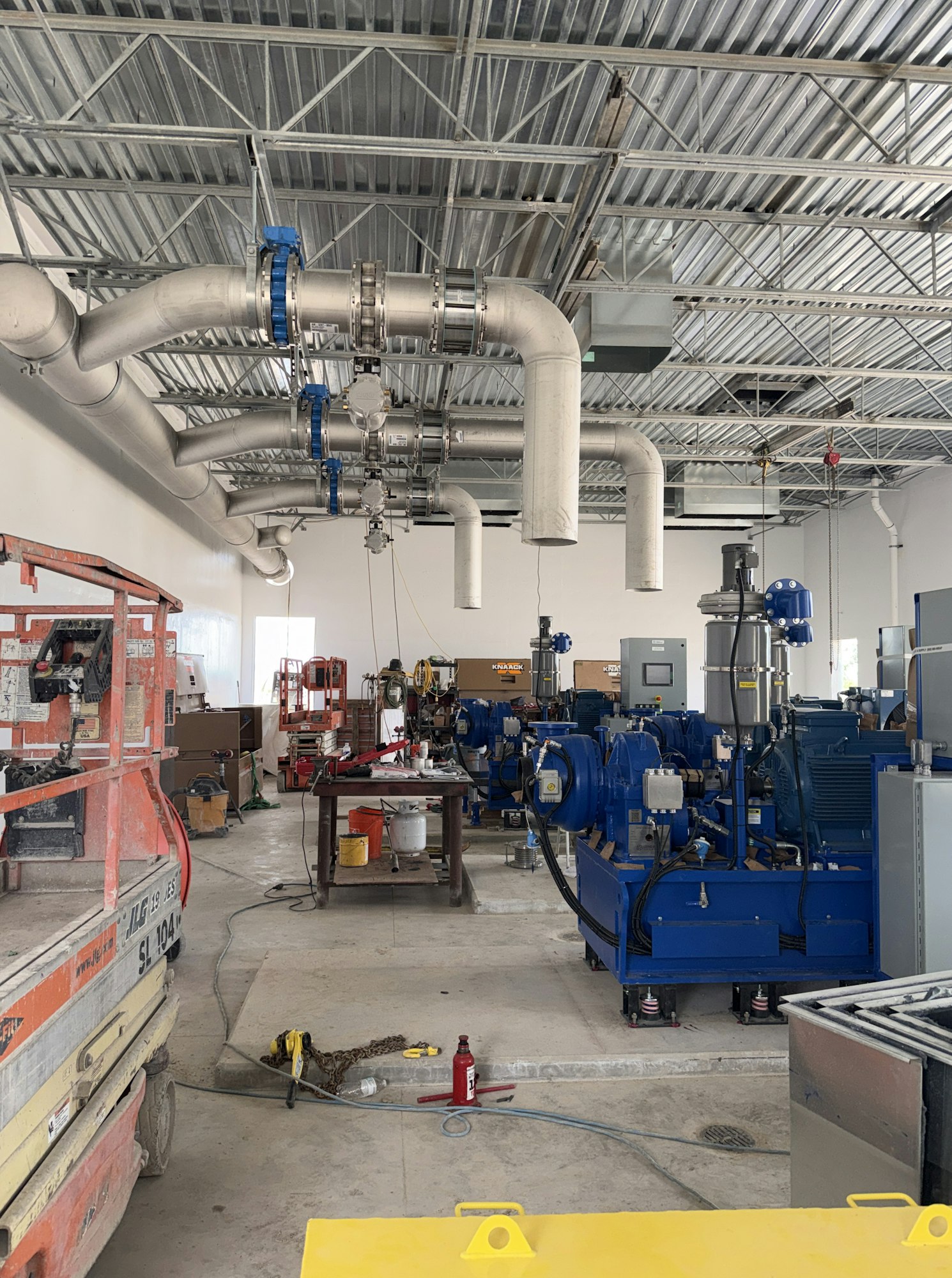 The image shows an industrial space with machinery, piping, and construction equipment, indicating ongoing installation or work.