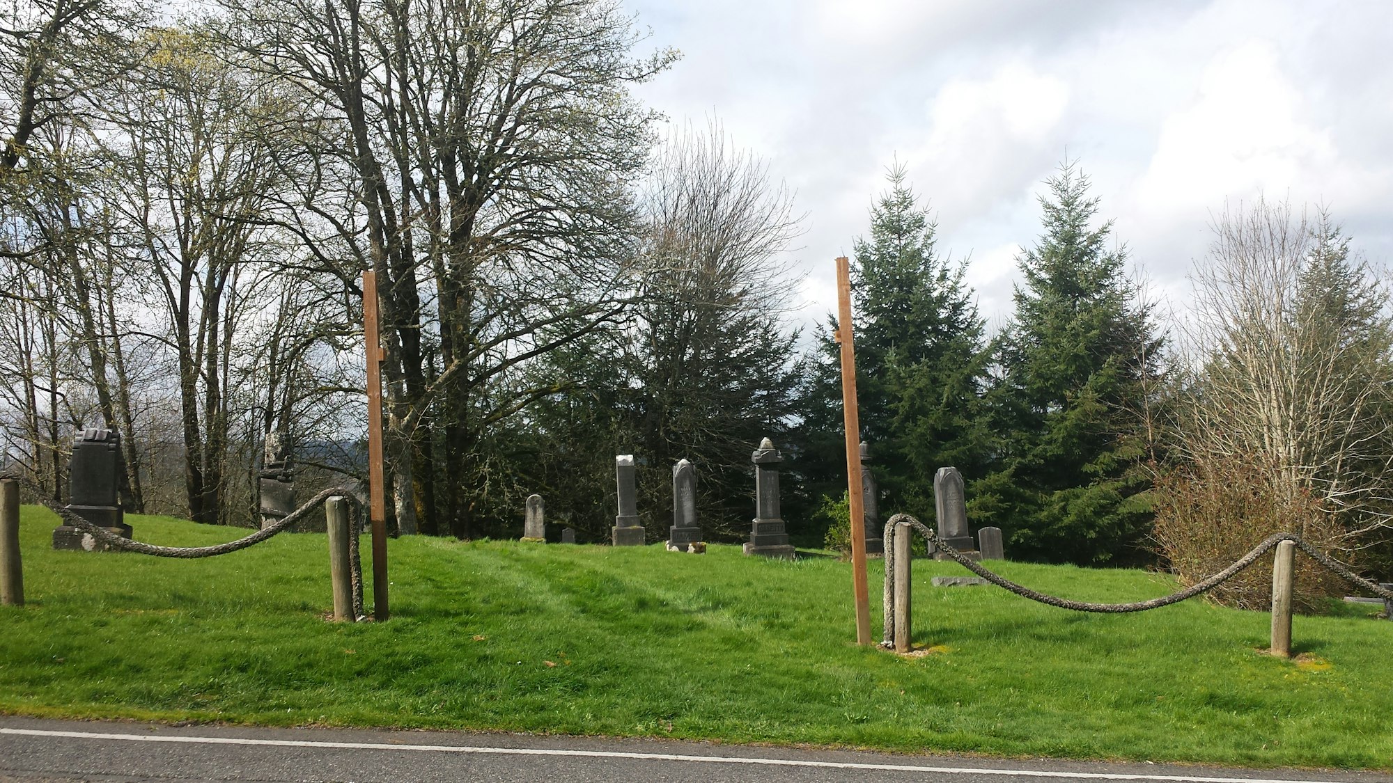 A grassy area with several gravestones, trees in the background, and a low fence made of wood and rope.