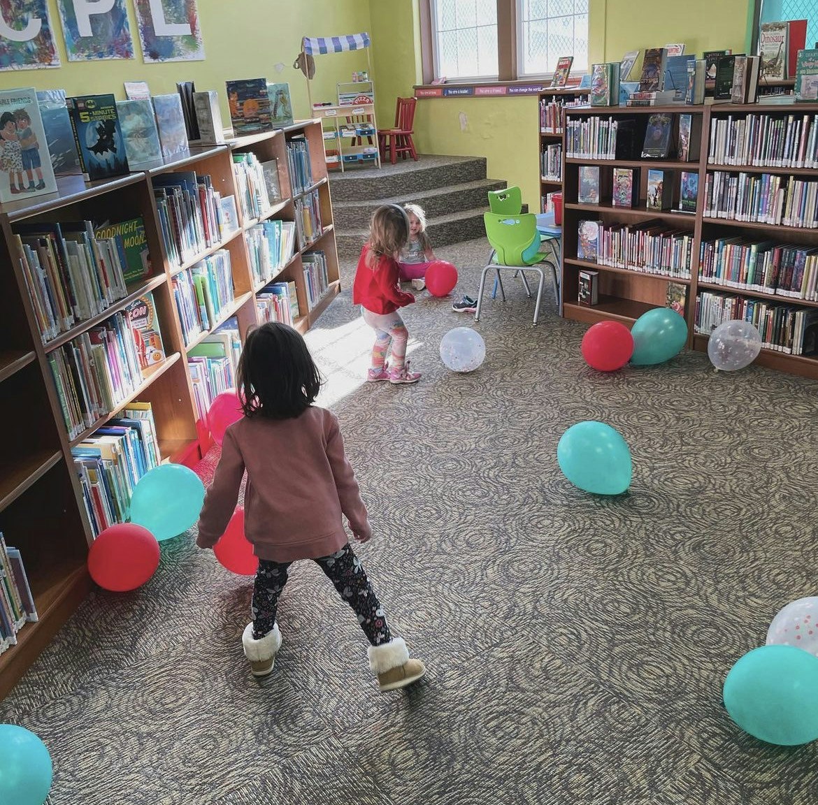 Two children play with colorful balloons in a library, surrounded by bookshelves and a cozy atmosphere.