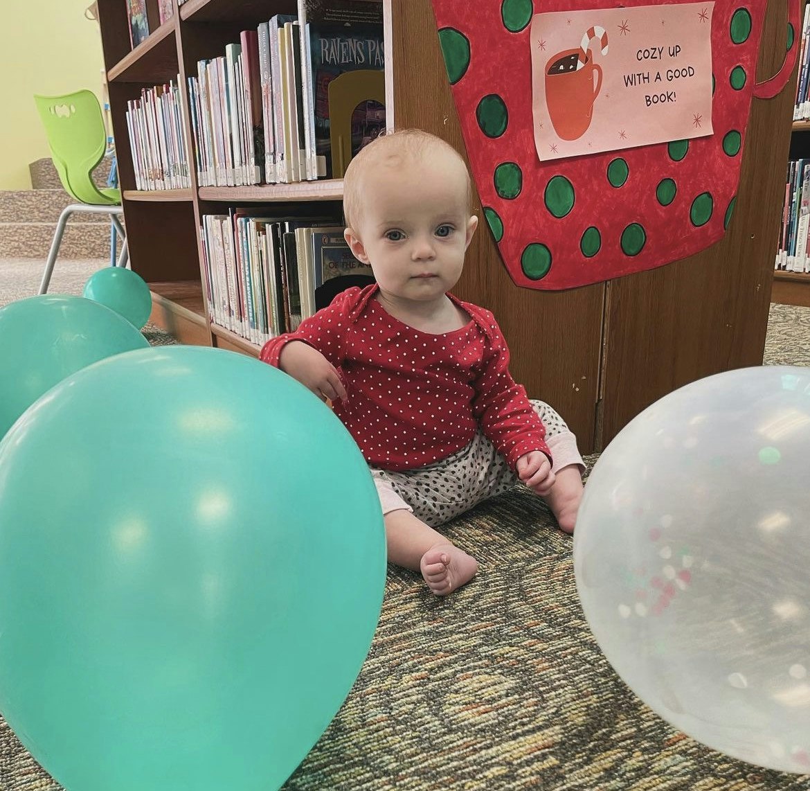 A baby sits amidst turquoise balloons in a library, with a cozy reading sign nearby.