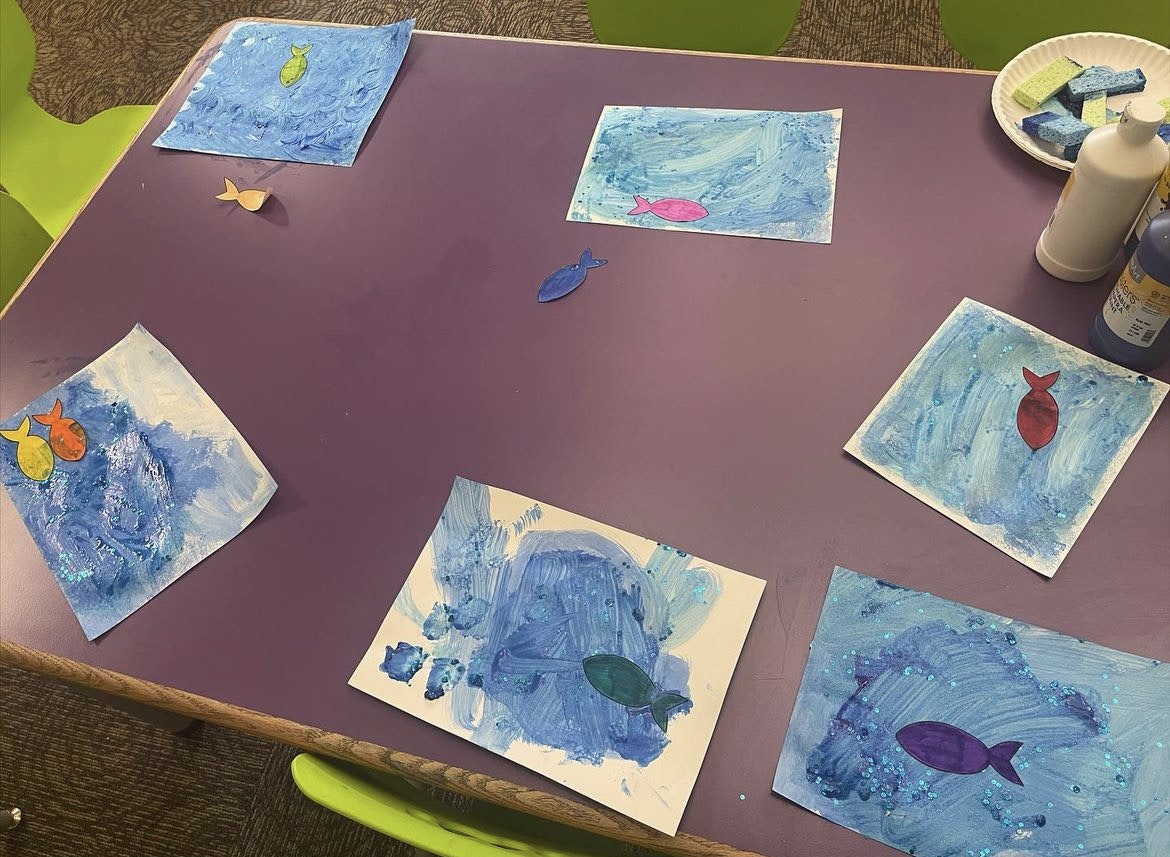 Colorful fish paintings on a purple table, with blue backgrounds and various craft supplies nearby.
