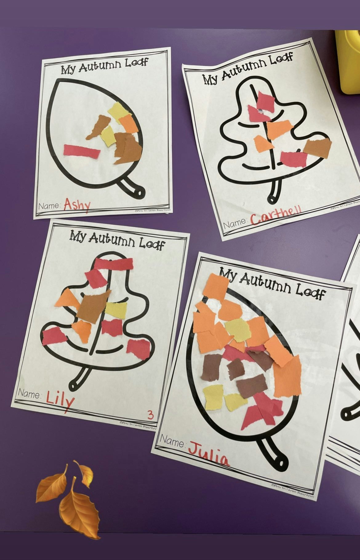 Colorful autumn leaf crafts made by children named Ashy, Carthell, Lily, and Julia, displayed on a purple table.