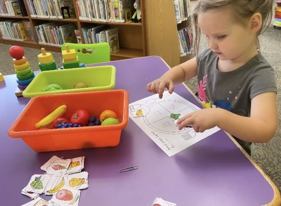 A young child plays with toy food and sorting cards at a library, engaging in hands-on learning activities.