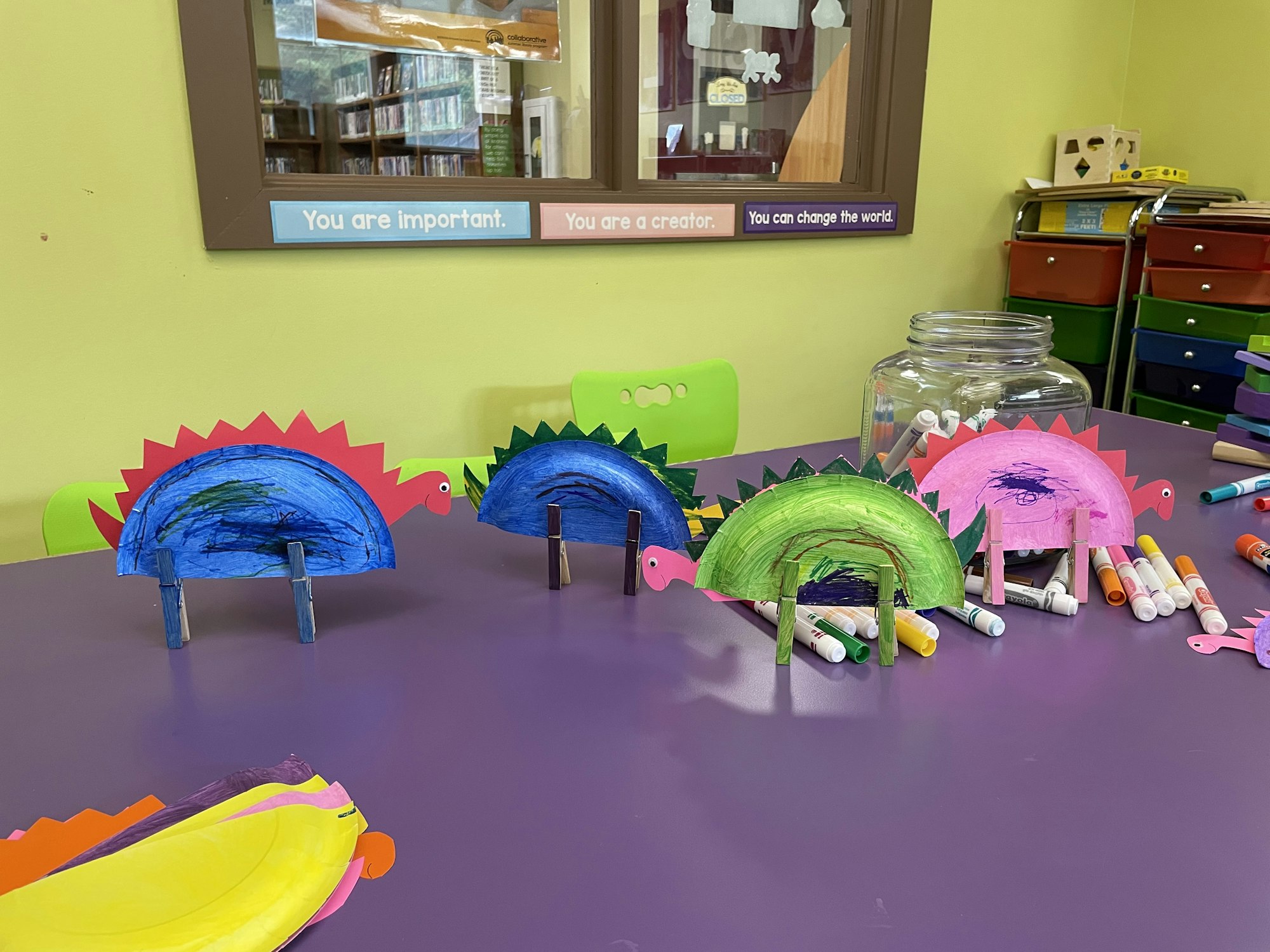 The image shows colorful paper dinosaur crafts on a table, surrounded by art supplies and a jar filled with markers.