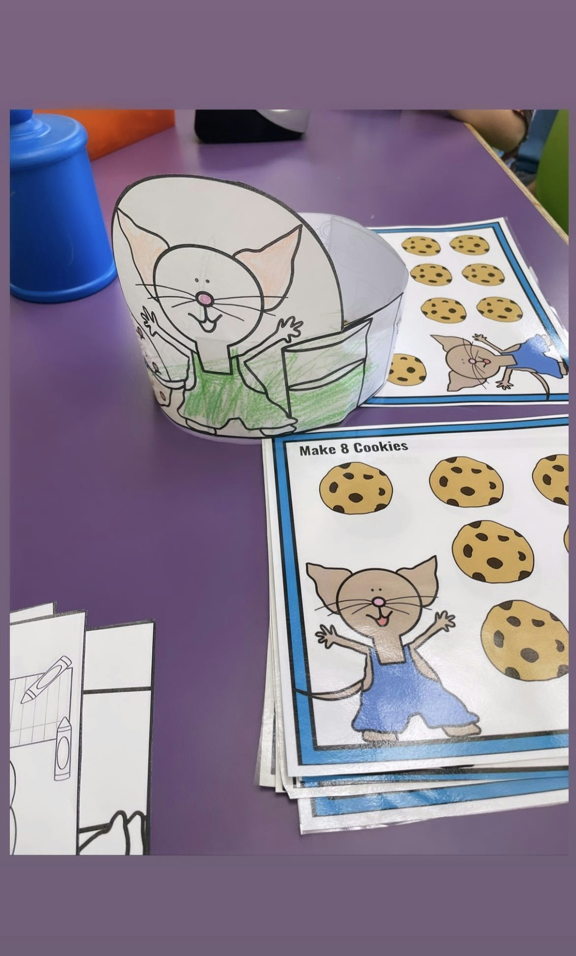 A colorful paper cat hat and worksheets featuring a cat making cookies, on a purple table.