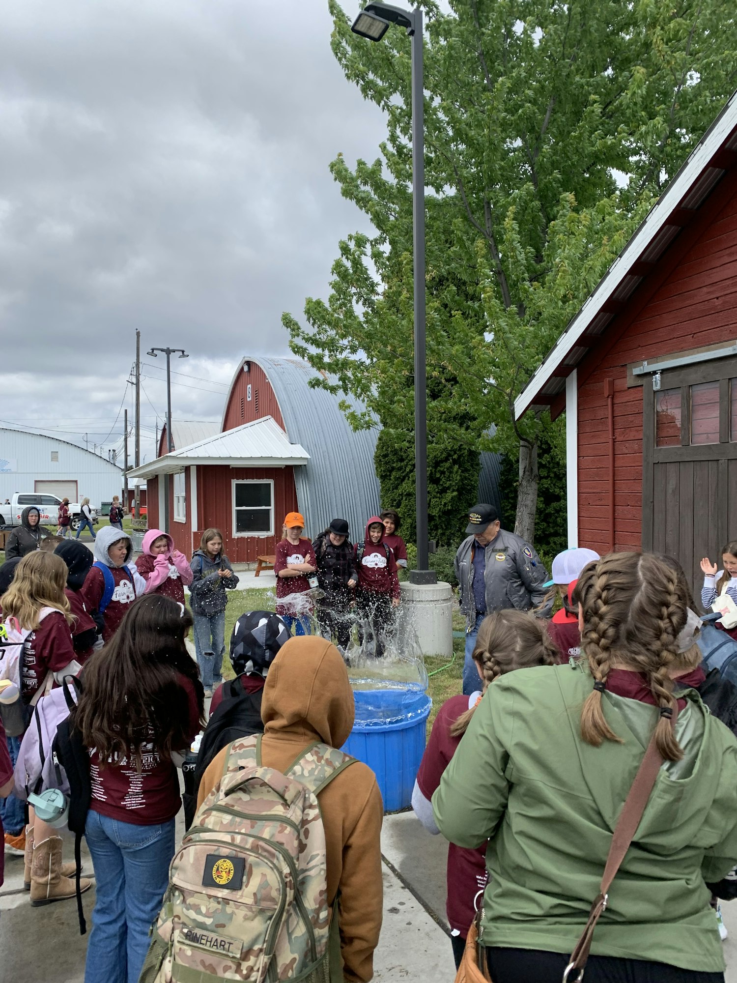 A group of people gathered around an outdoor area with a red building and trees in the background.