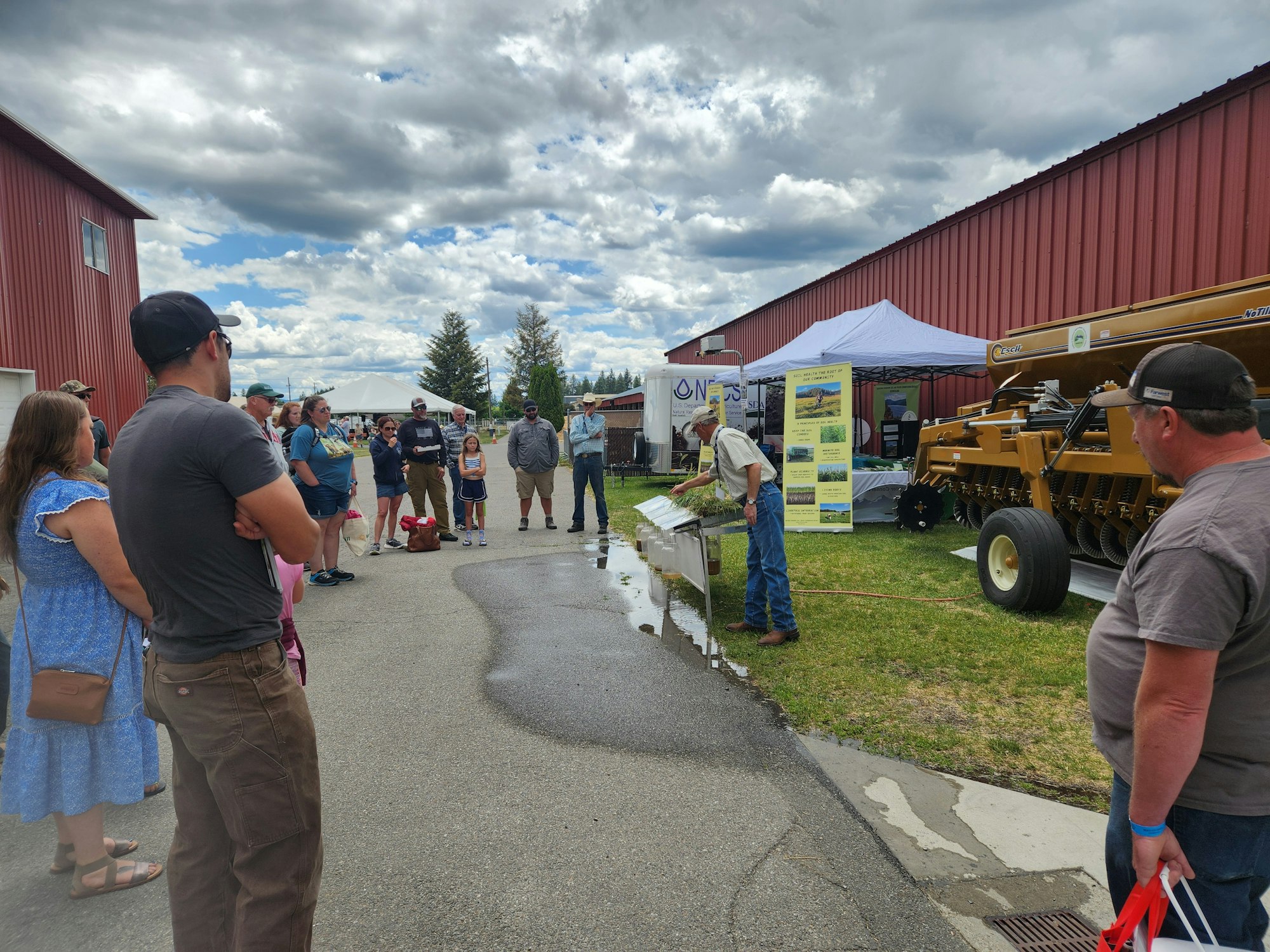 A group of people outdoors near a red building, watching a demonstration with farming equipment and a tent in the background.