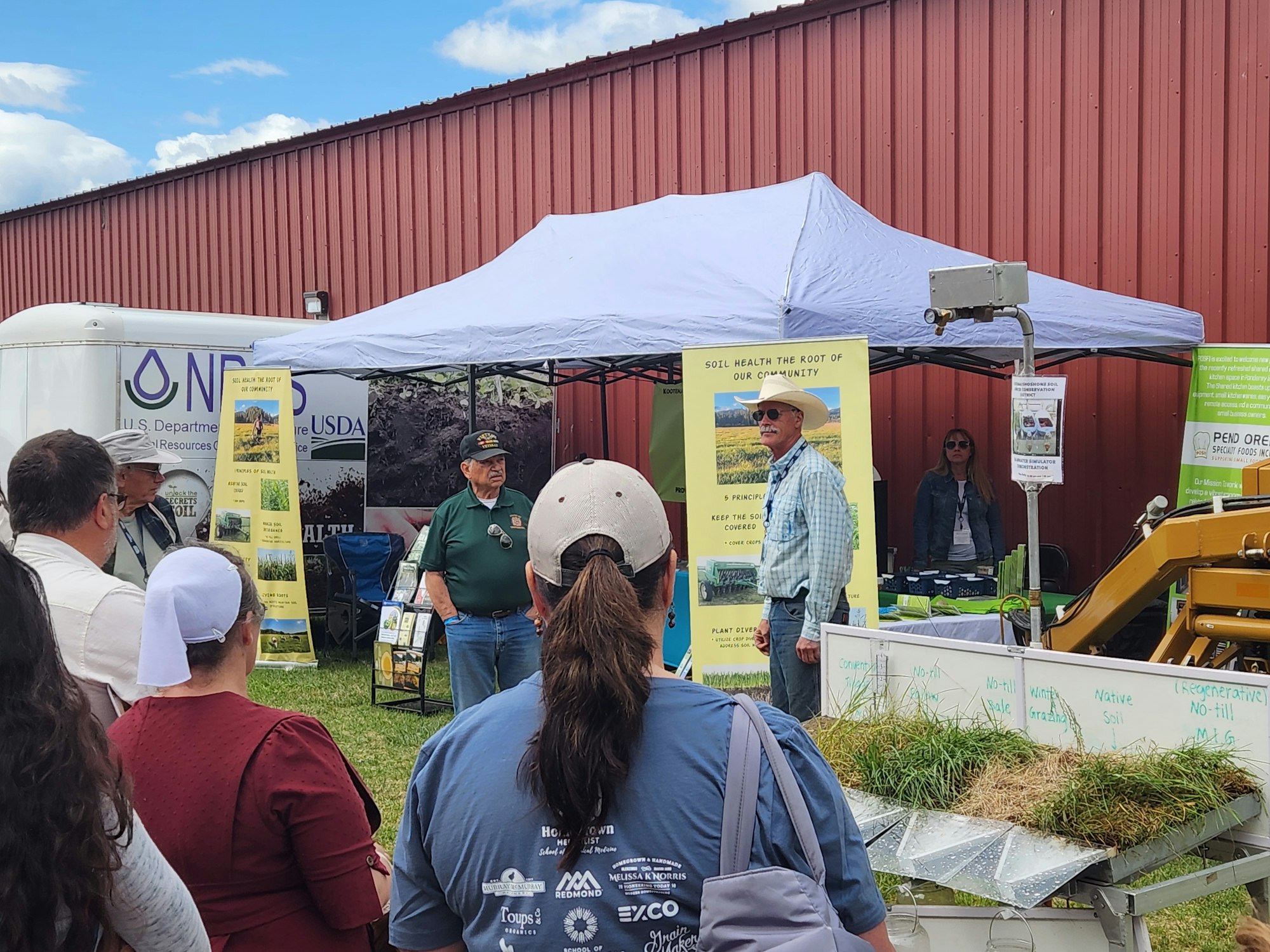 People listening to a presentation about soil health at an outdoor exhibit with informational posters and a small model display.