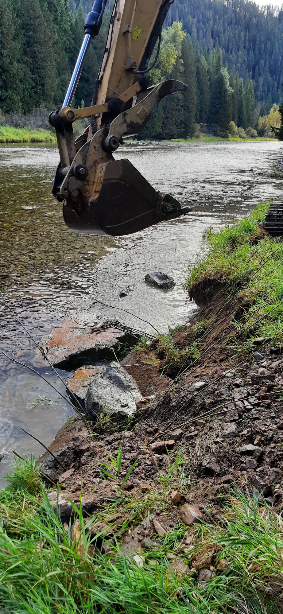 An excavator bucket near a waterbody, trees in the background; image is rotated 90 degrees.