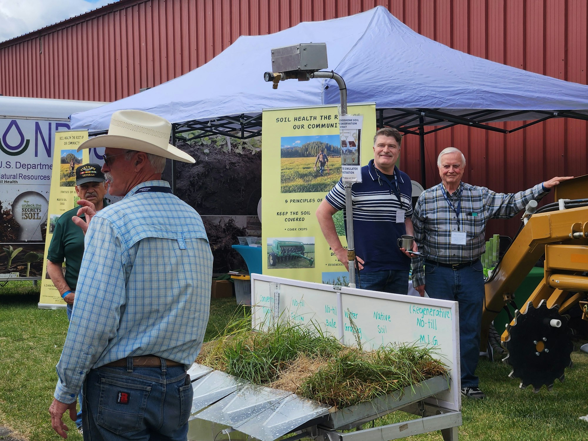 A group of men at an agricultural booth with soil and plant display, discussing soil health and conservation practices.