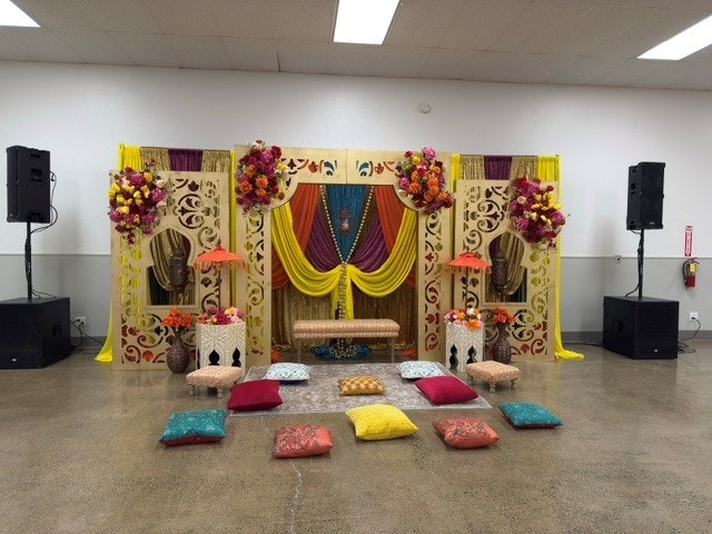 A vibrant event setup featuring colorful fabric drapes, decorative flower arrangements, a seating area with cushions, and audio equipment.