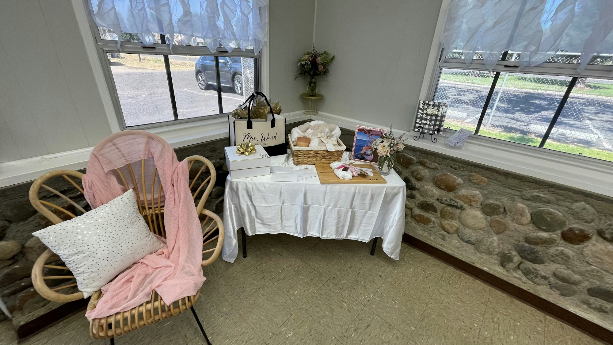 A cozy corner with a wicker chair, cushions, a table with gifts and flowers, near a window with sheer curtains.