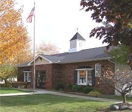 The image shows a brick building with a flagpole, surrounded by trees in fall foliage. It has a welcoming porch and classic architecture.