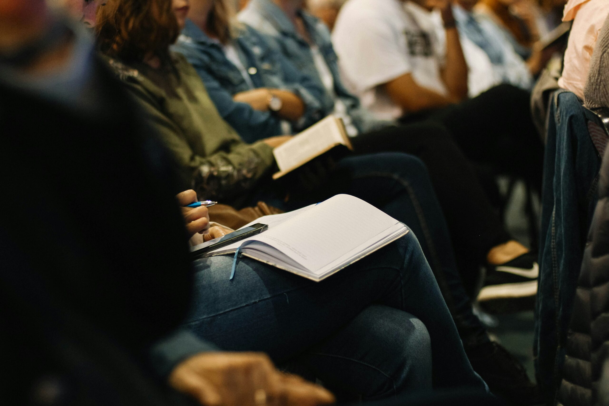 Audience seated with notebooks, focus on one person writing.