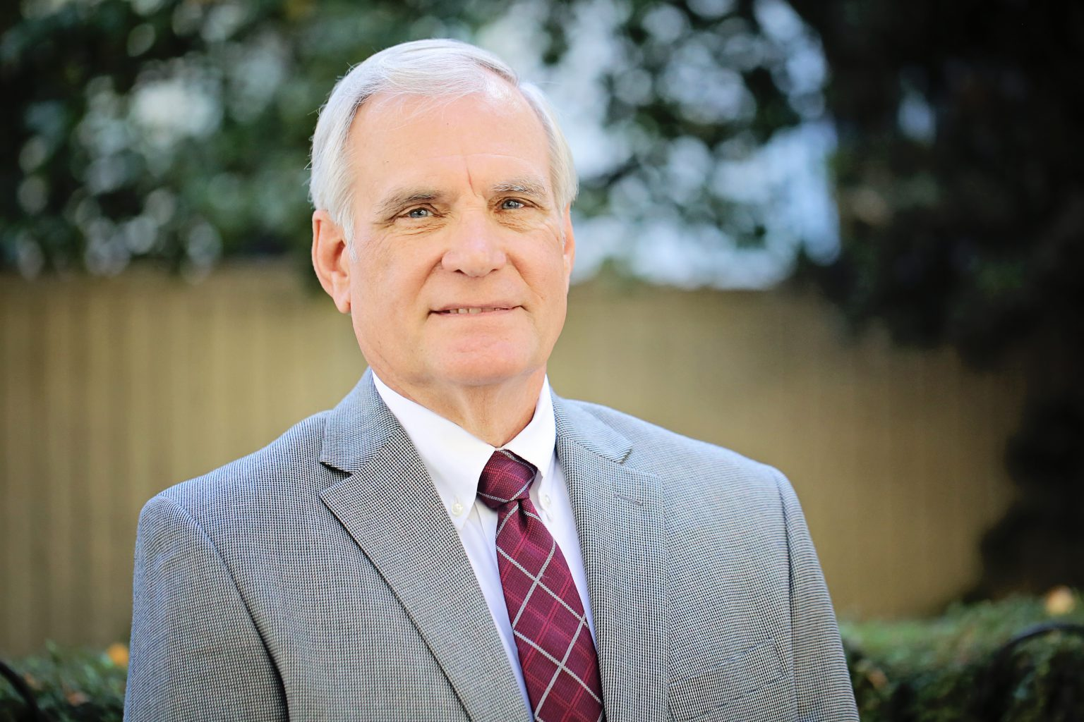 A portrait of a smiling man in a suit and striped tie, with a blurred background of foliage and a fence.