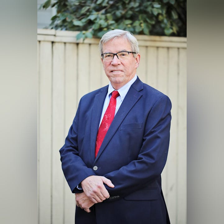 Man in suit with red tie standing by a fence; appears professional.