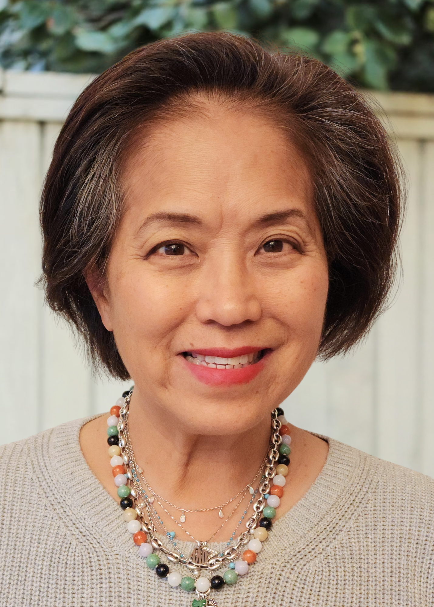 A woman smiles, wearing a cozy sweater and layered necklaces, in a green outdoor setting with a blurred background.