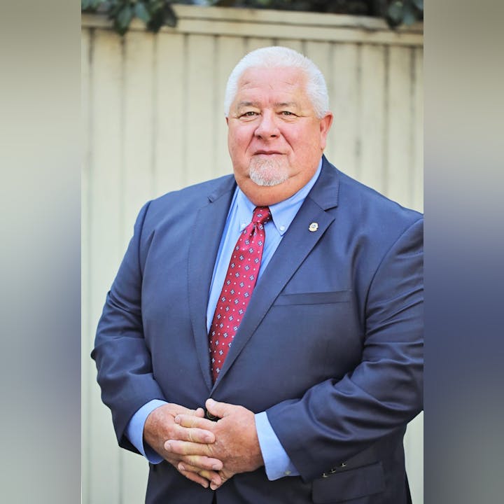 A man in a suit with a lapel pin, wearing a red tie, standing in front of a fence.