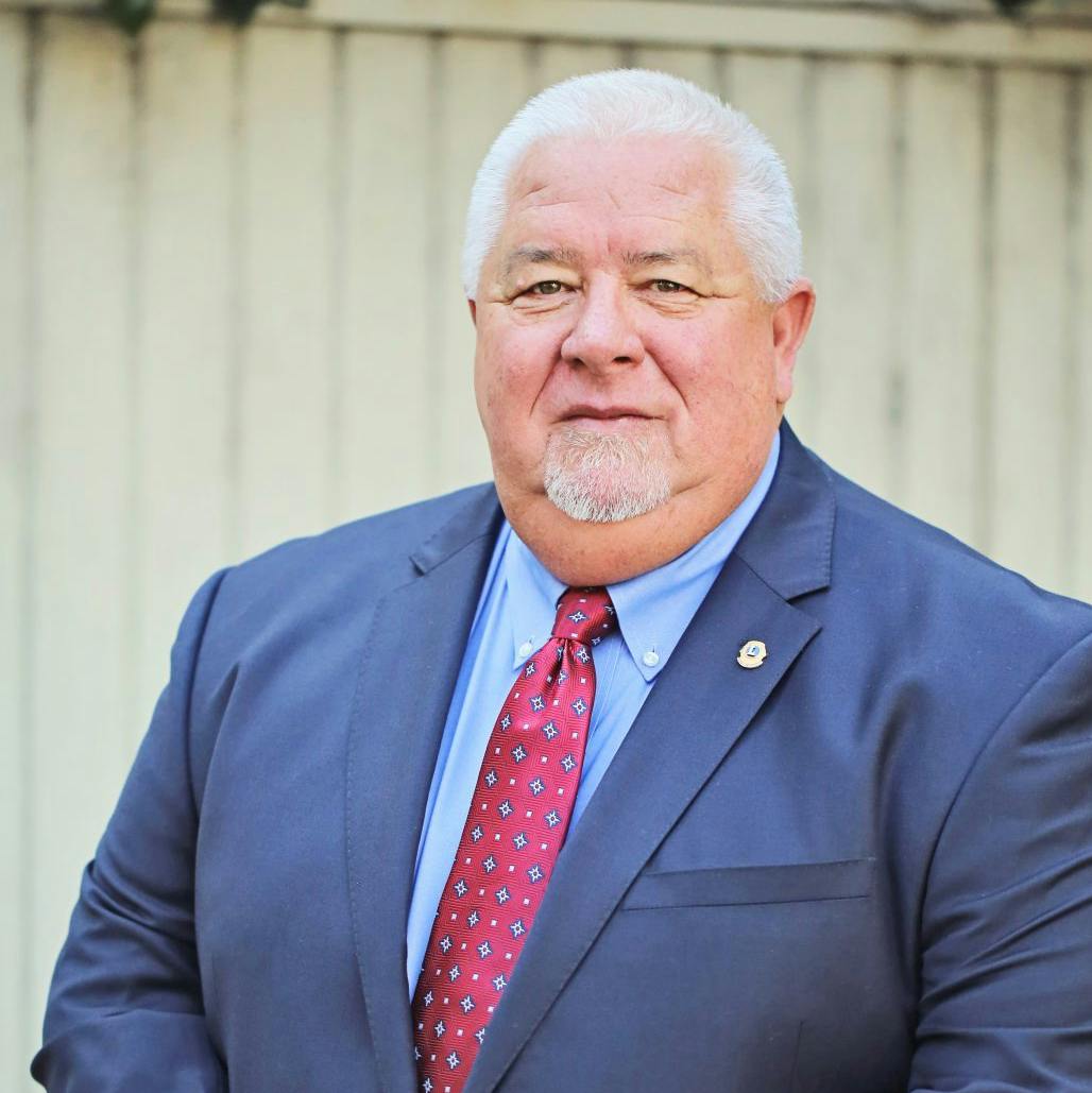 A man in a suit with a lapel pin, wearing a red tie, standing in front of a fence.