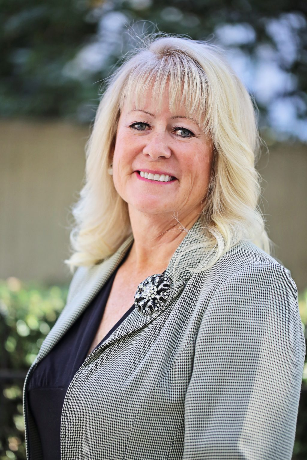 A smiling woman with blonde hair, wearing a patterned blazer and a decorative brooch.