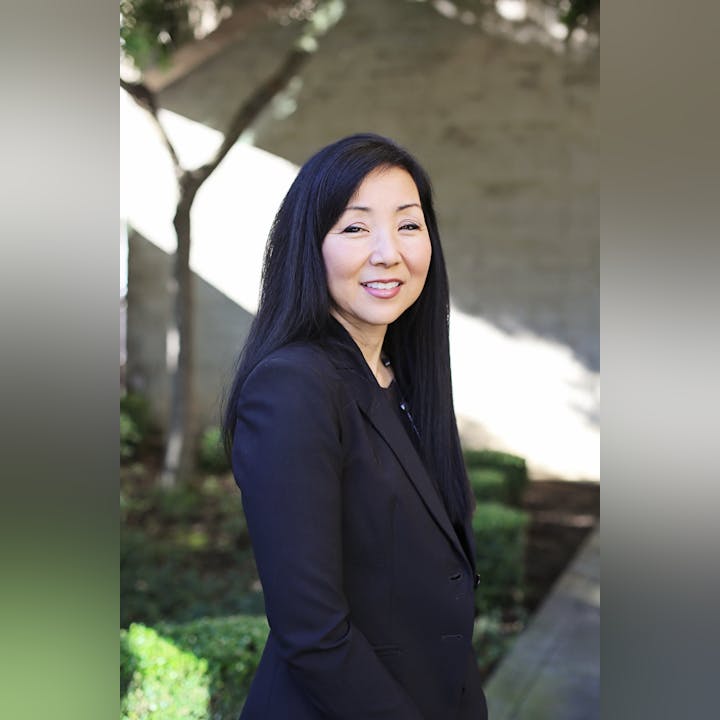 A person in a black blazer stands outside, smiling, with trees and greenery in the background.