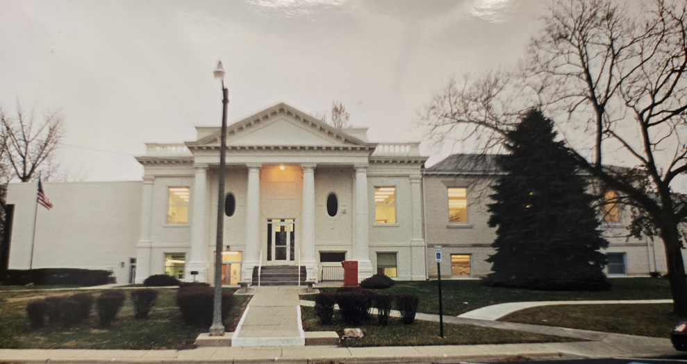 A large, classic-style building with columns and an American flag nearby, framed by trees.