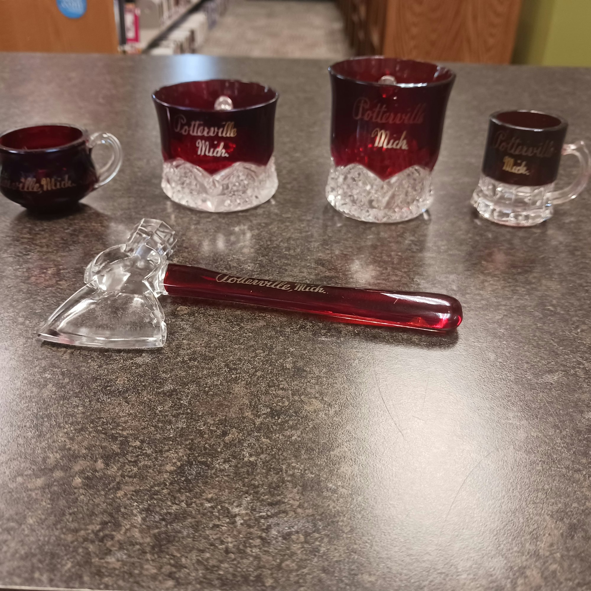 Four red and clear glass cups and a glass axe-shaped object with "Potterville Mich." engraved on them, placed on a table.