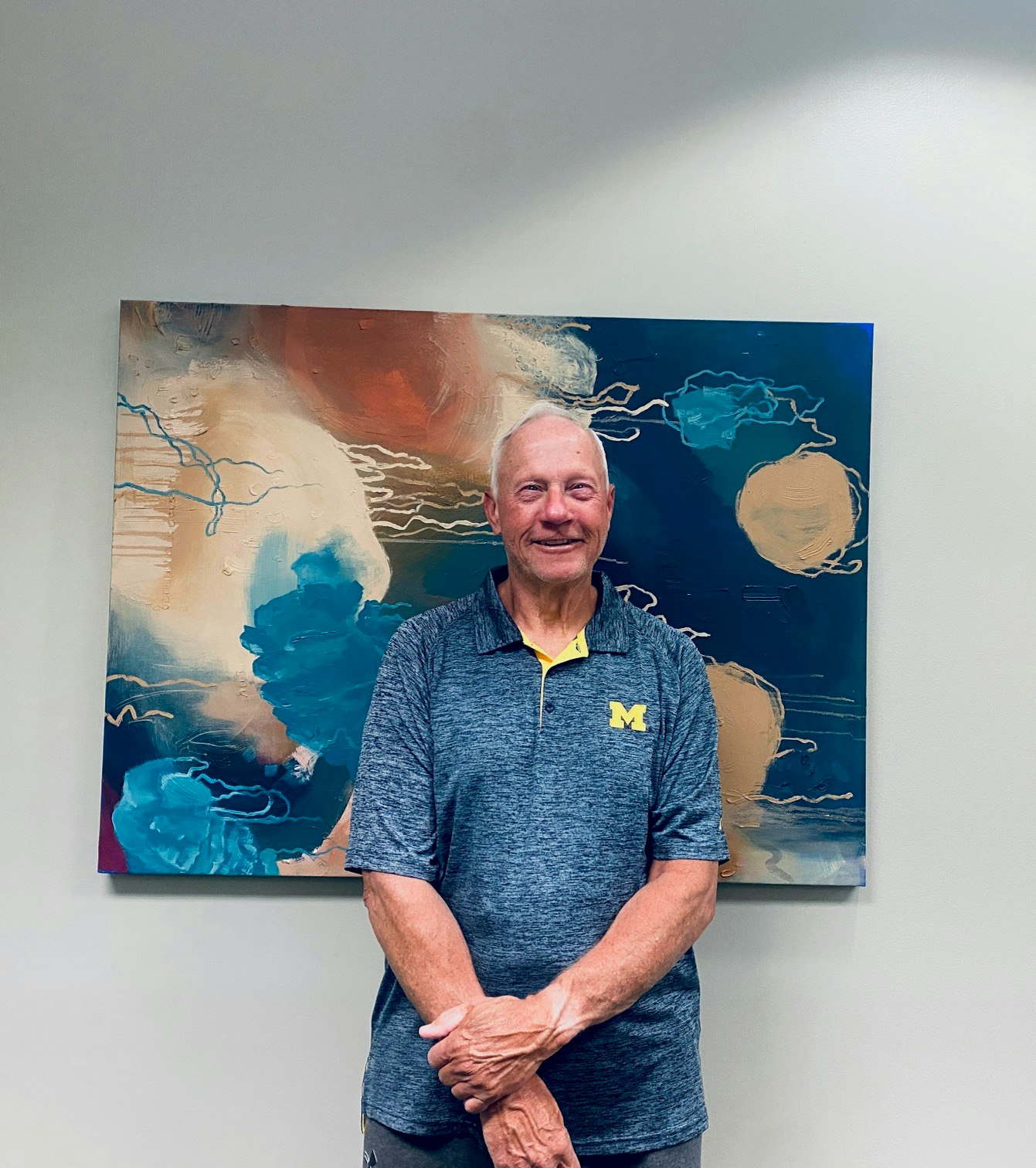 An older man stands smiling in front of an abstract painting with blue and warm tones. He's wearing a shirt with a university logo.