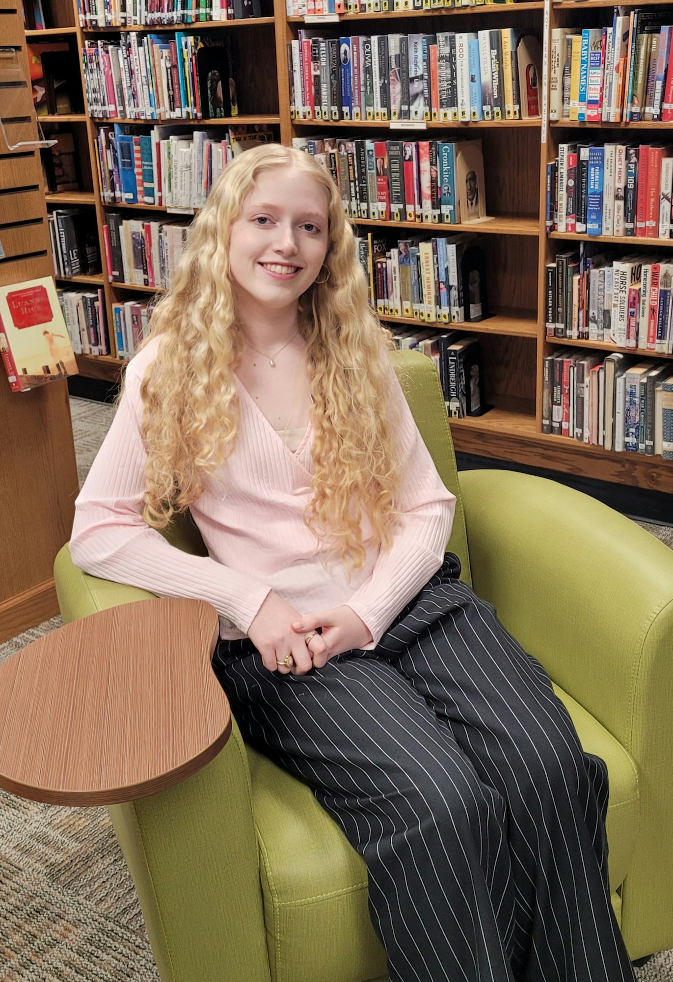 A person smiling, sitting in a green chair by bookshelves in a library setting.