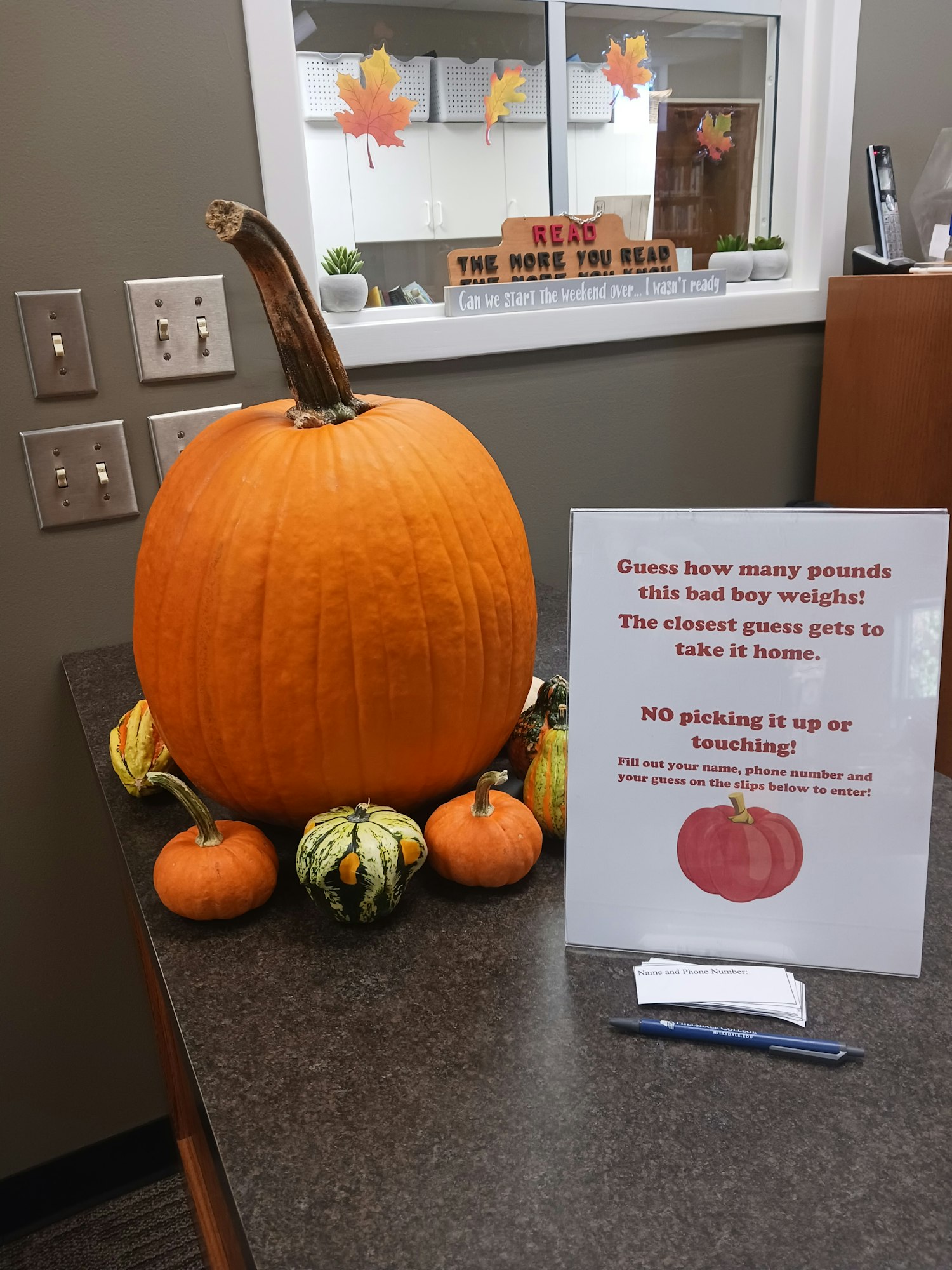 Large pumpkin, gourds, guessing game sign, pen, cards on a counter. Autumn theme. Image rotated 90 degrees clockwise.