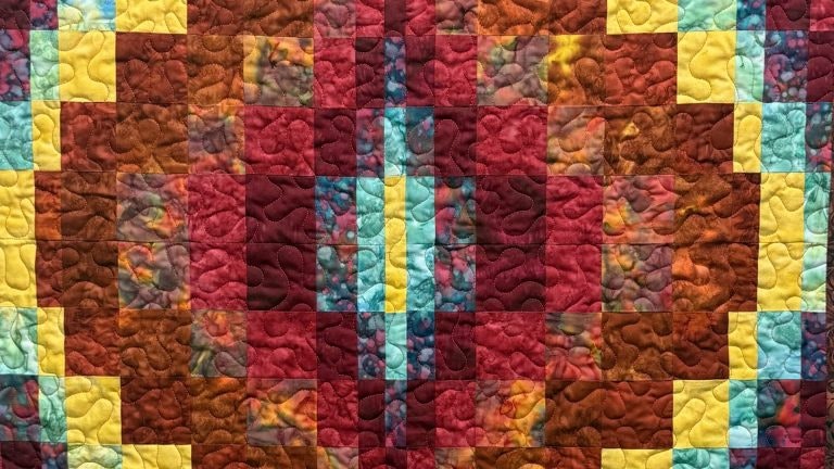 The image shows a colorful quilt featuring geometric patterns in warm colors like red, orange, and yellow with contrasting blue accents.
