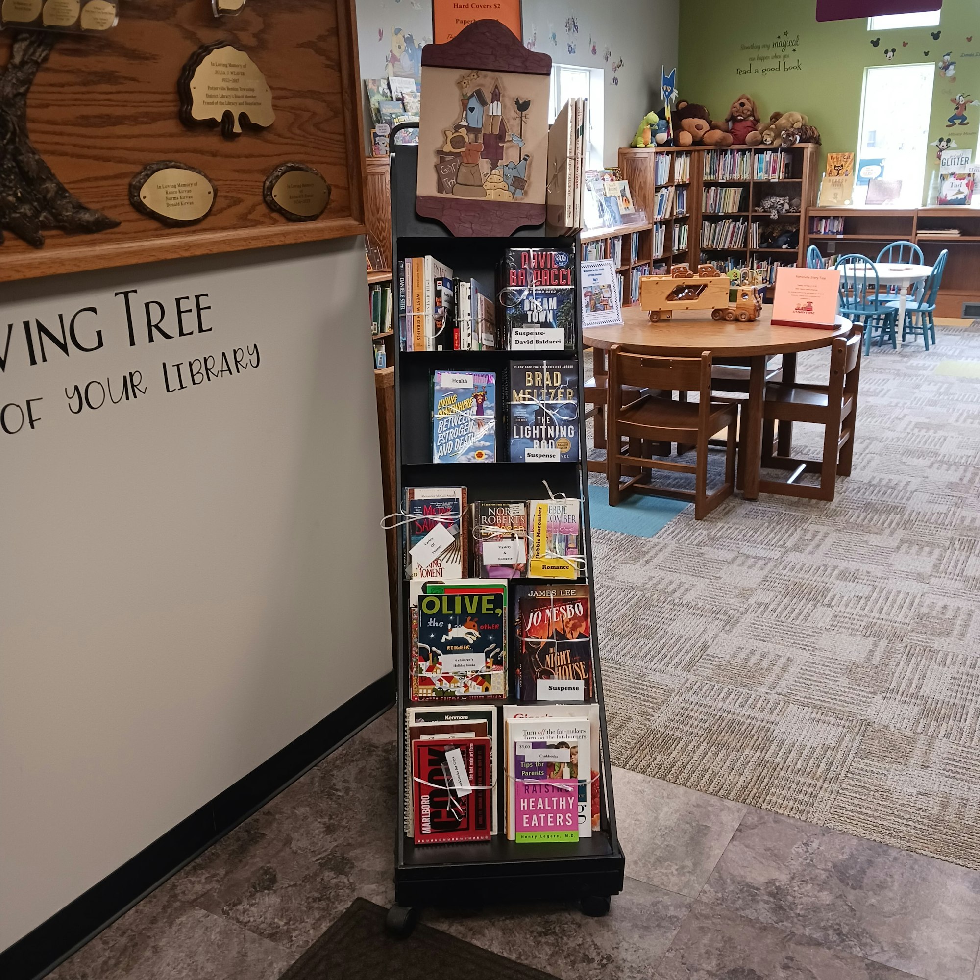 Library corner with bookshelves, tables, chairs, and a display rack with various books.