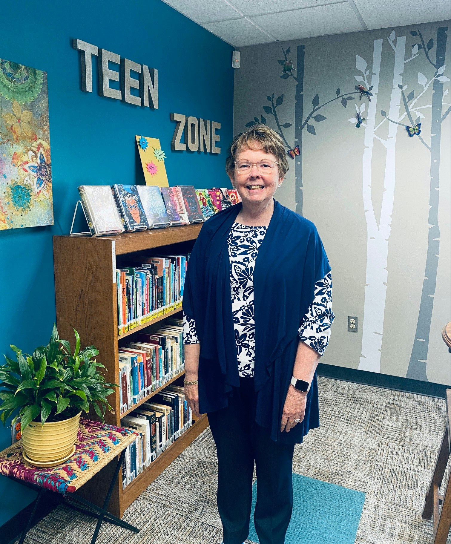 A smiling woman stands in a "Teen Zone" library area with bookshelves, a plant, and decorative wall art around her.