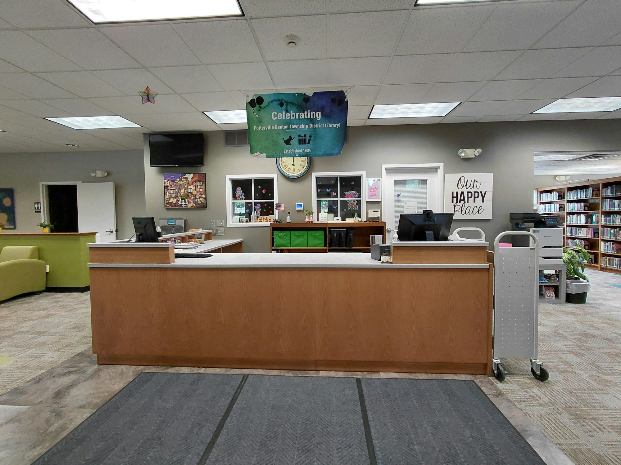 A library reception area with a counter, computers, colorful decor, and shelves filled with books in the background.