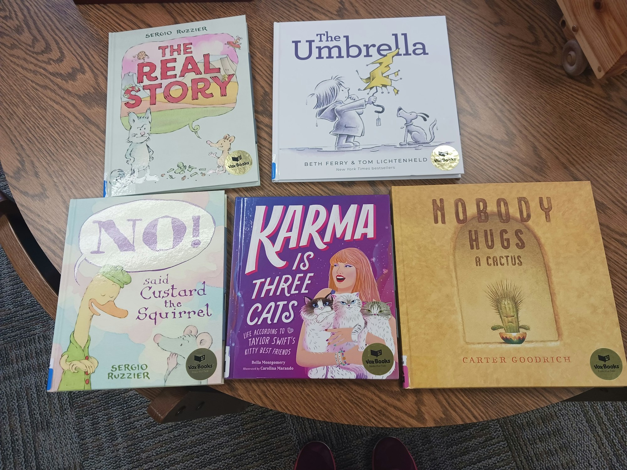 Five children's books on a table: "The Real Story," "The Umbrella," "NO! Said Custard the Squirrel," "Karma is Three Cats," and "Nobody Hugs a Cactus."