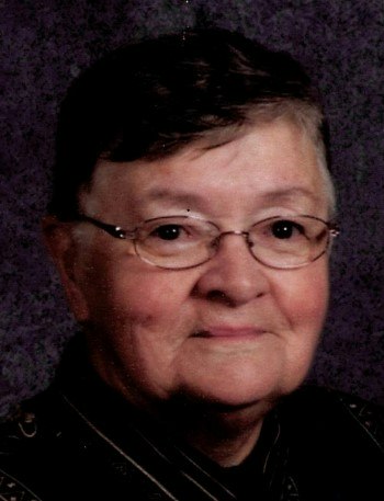 A close-up portrait of an older woman with short hair and glasses, smiling gently against a dark background.