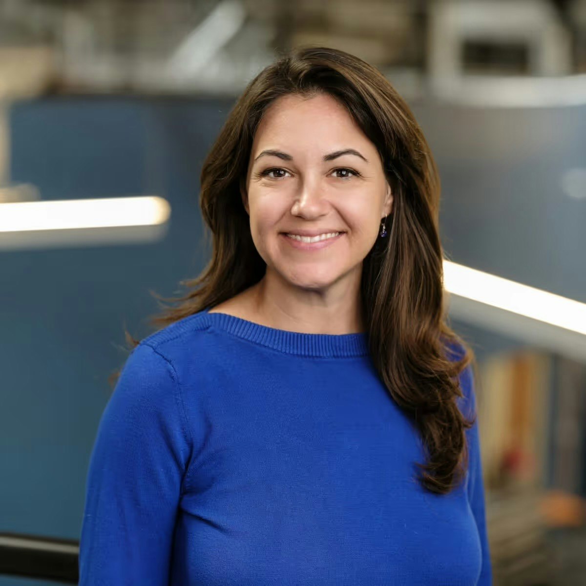 A person in a blue sweater smiling indoors with a blurred background.