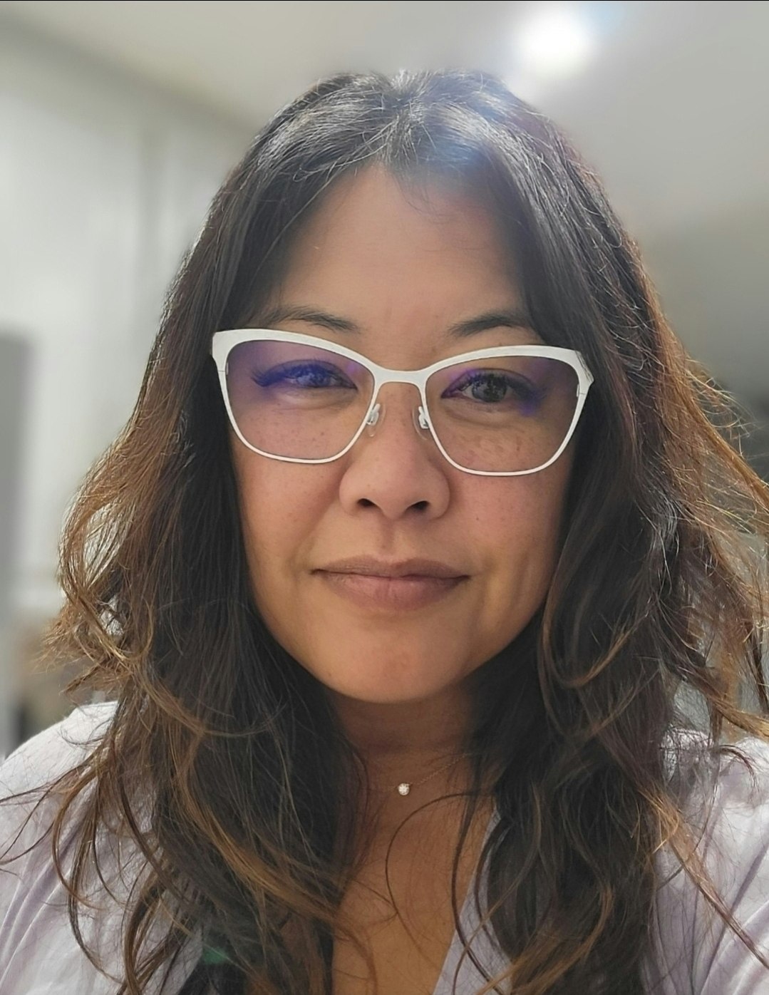 A person with long hair and glasses, dressed in white, smiling subtly.