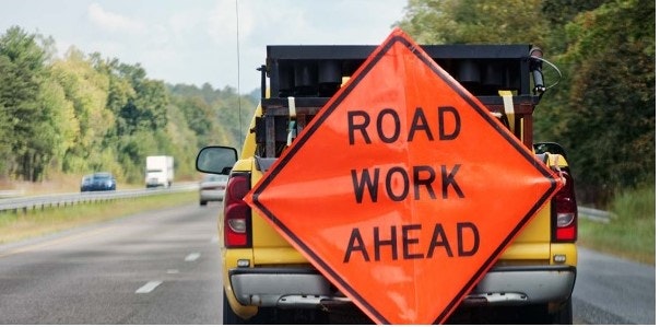 A bright orange road sign warns drivers of upcoming road work ahead. A truck displays the sign on a highway.
