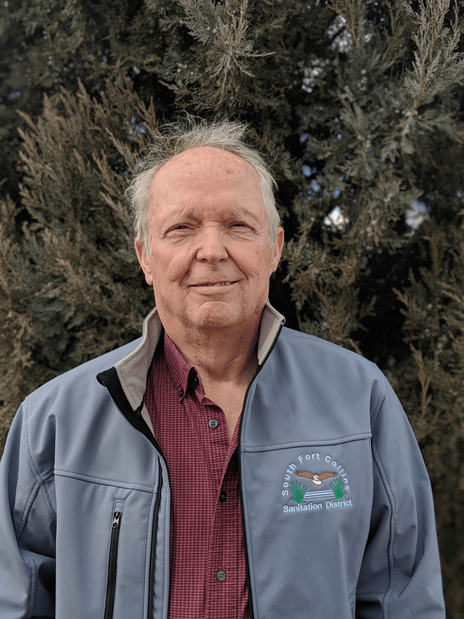 A man in a jacket with "South Fort Collins Sanitation District" logo, standing before evergreen trees.