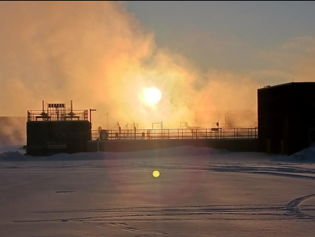 Sunrise and mist over a snowy industrial scene with buildings and ice-covered ground.