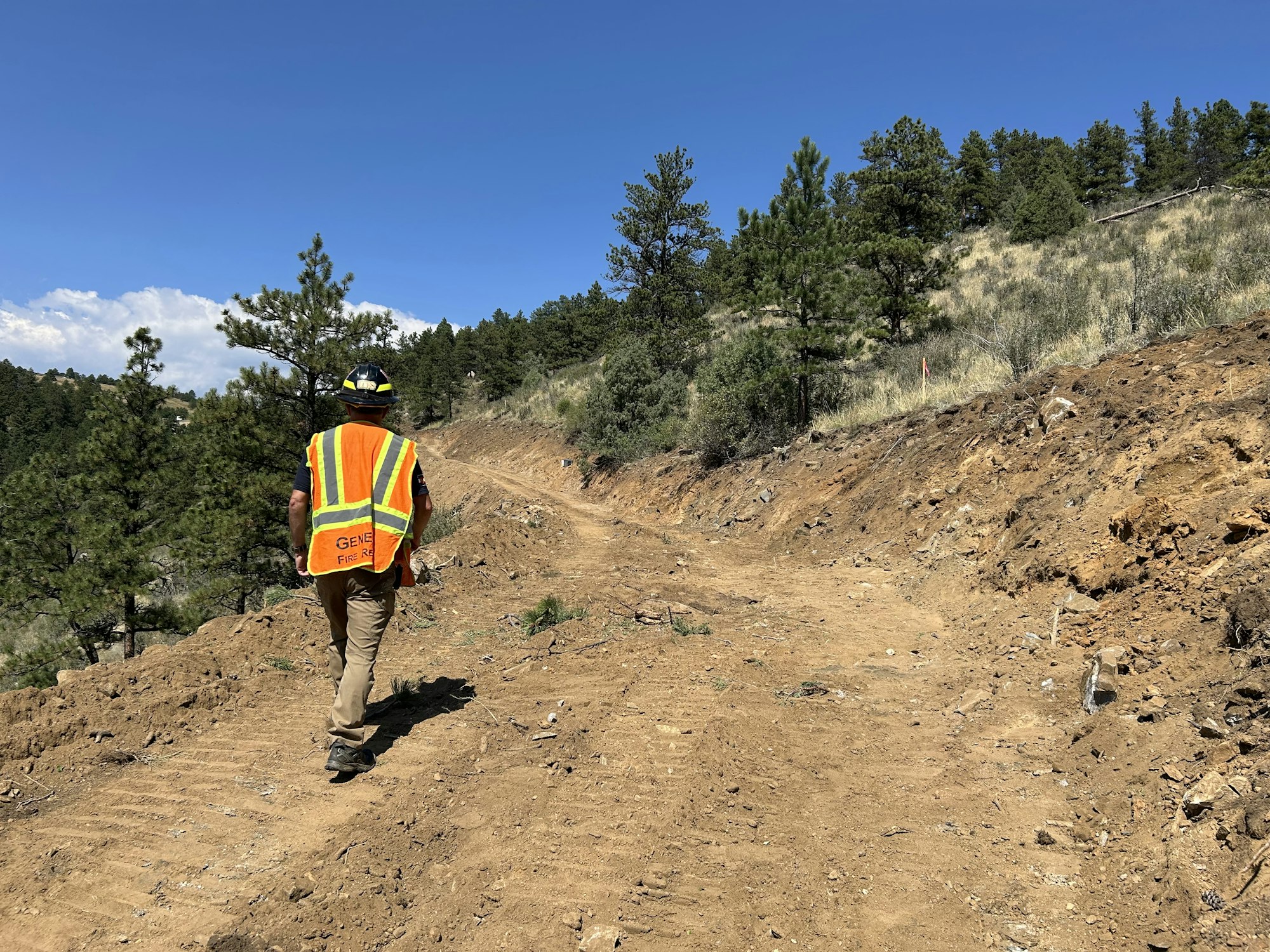 A person in a high-visibility vest and helmet walks on a dirt trail surrounded by trees and a clear blue sky.