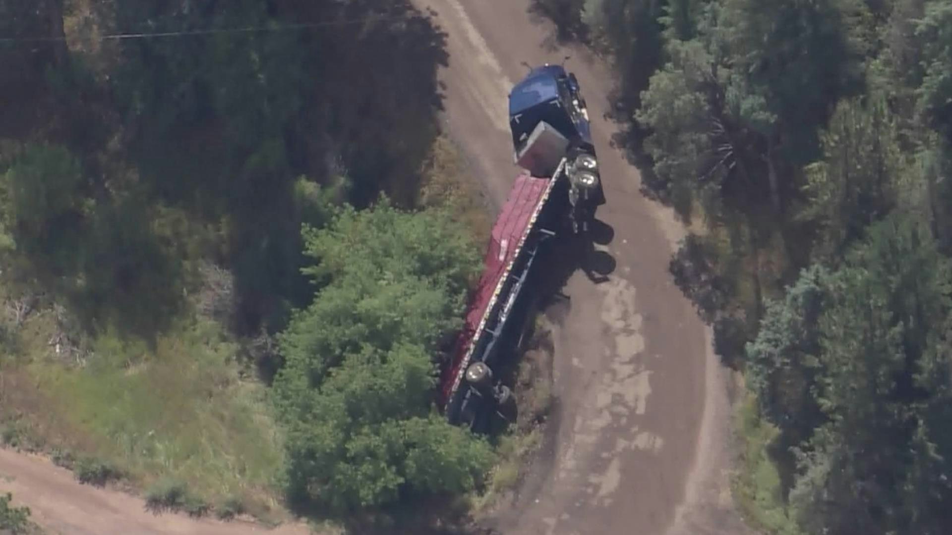 Aerial view of a truck accident with a trailer overturned on a dirt road, surrounded by trees.