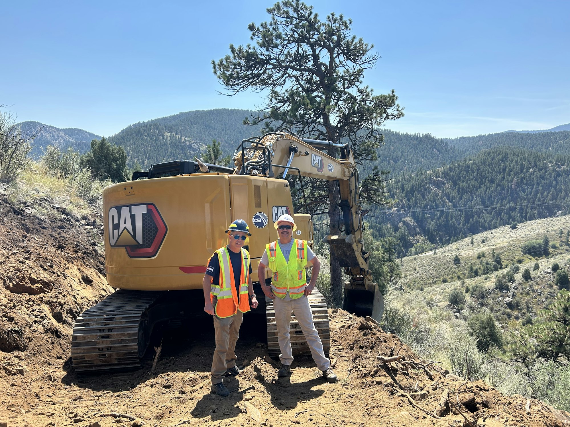 Two workers in safety gear stand beside a CAT excavator on a hilly landscape with trees and mountains in the background.