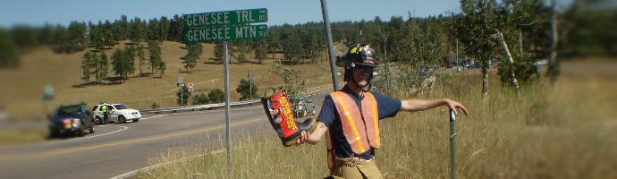 A person in a safety vest and hard hat holding a sign by a road with a "Genesee TRL" sign in the background.